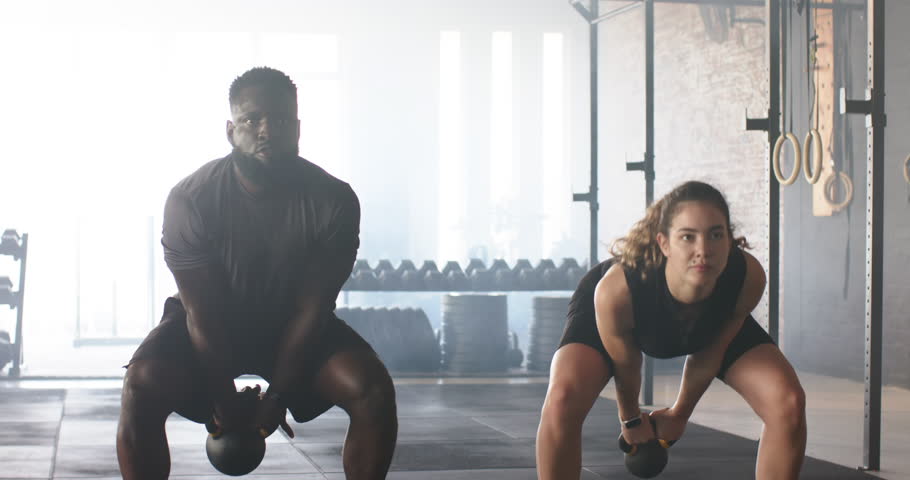 African American male, white female hinging hips swinging kettlebells overhead for training in gym. Strength, fitness, workout, dynamic, power, gym, equipment