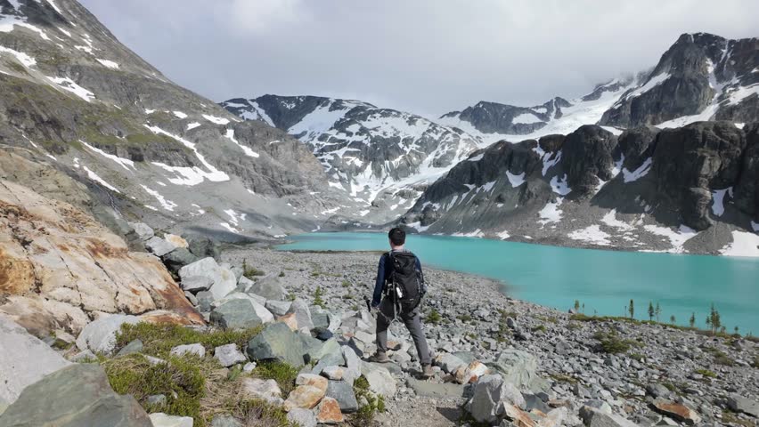 Man Hikes Towards Vibrant Turquoise Glacier Lake Amidst Majestic Snow-Capped Mountains in British Columbia, Canada