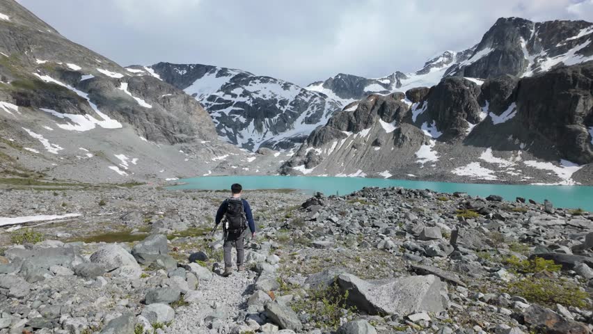 Man Hiking Towards a Stunning Turquoise Lake Surrounded by Majestic Snow-Capped Mountains in British Columbia.