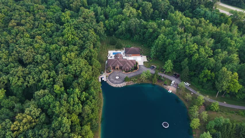 4K aerial drone footage of a private countryside house near a scenic lake, surrounded by green trees and peaceful nature. Beautiful cinematic flyover of rural property and landscape.