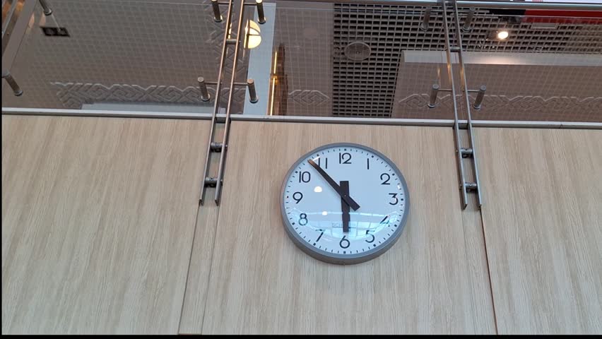 A modern round classic analog clock with a white face, black numbers and hands hangs on a wooden wall. The wall is part of an interior space with a glass ceiling and metal railings. The scene takes pl