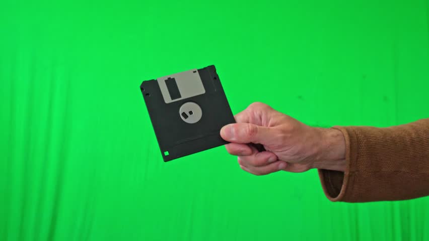 A nostalgic and technical mood is captured as a hand holds a classic 3.5-inch floppy disk, ready for integration into various digital or historical themes against a vibrant green screen - Powered by Shutterstock - Get 15% off with code: PIKWIZARD15