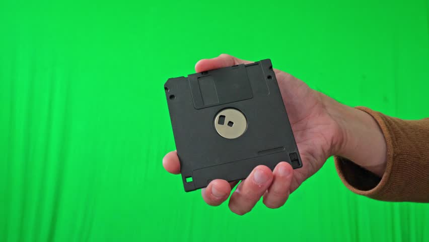 A nostalgic and technical mood is captured as a hand holds a classic 3.5-inch floppy disk, ready for integration into various digital or historical themes against a vibrant green screen - Powered by Shutterstock - Get 15% off with code: PIKWIZARD15