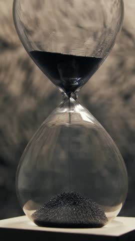 Clear glass hourglass with fine black sand steadily falling symbolising passing time and looming deadline