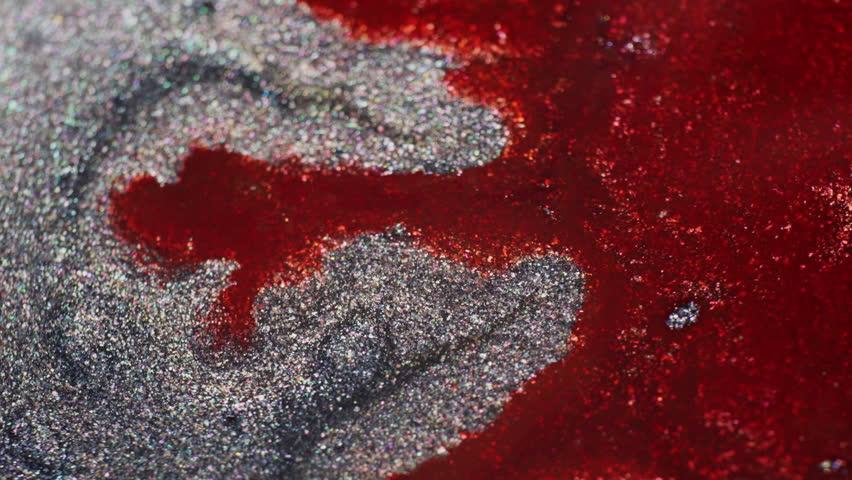 Macro shot showing glittery grey and orange ink reacting to added water. The red ink spreads and mixes with the grey, forming mesmerizing, fluid patterns.