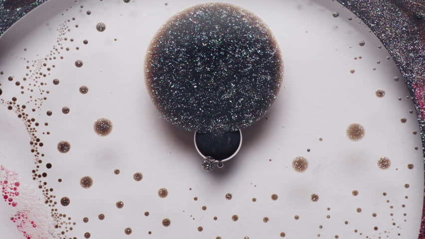 Macro view of vibrating black glitter ink in a white liquid, surrounded by red and white ink droplets. The ink pulses and swirls intensely.