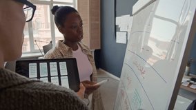 Black female team lead presenting at whiteboard planning timeline for UI design project with colleague holding laptop with interface mockups for mobile app while collaborating at office, slow motion - Powered by Shutterstock - Get 15% off with code: PIKWIZARD15