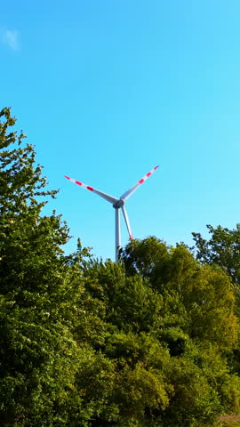 Rotating blades of windmill above the lush greenery of the trees. Low angle view at the wind turbine. Blue sky at backdrop. Vertical video.