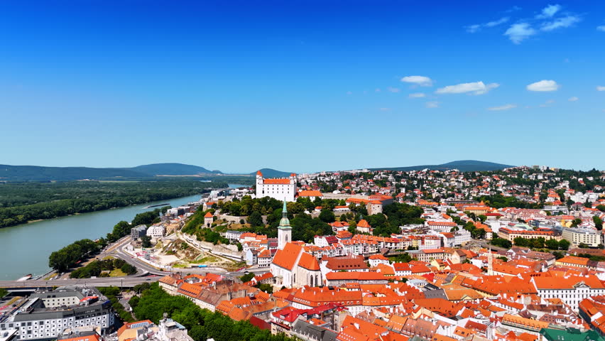 Sun lighting the orange roofs of the old-fashioned historical buildings in the cityscape of Bratislava, Slovakia. Waterscape of the Danube river and green hills at backdrop. Aerial view.
