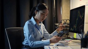 Young businesswoman analyzing financial charts displayed on a computer screen and smartphone, pointing at the monitor with a pen while sitting at her desk in a dimly lit office - Powered by Shutterstock - Get 15% off with code: PIKWIZARD15