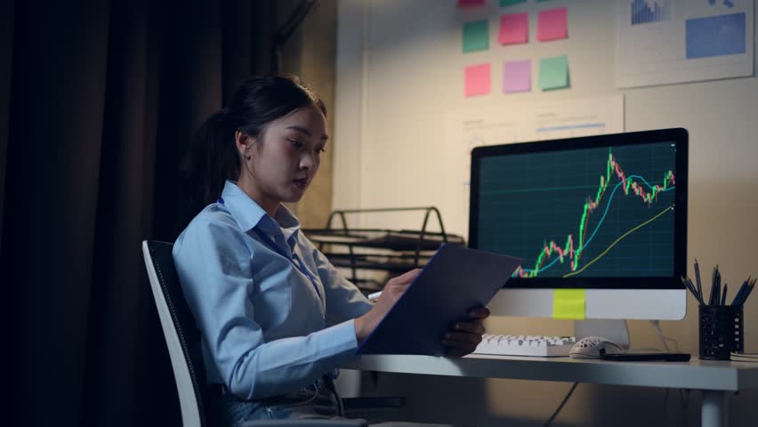 Young professional businesswoman carefully analyzing financial data on computer screen, taking detailed notes on clipboard while working late at modern office desk during night hours