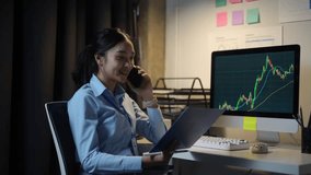 Young Asian businesswoman analyzing financial charts while talking on phone, working late at night, displaying professional focus and corporate dedication from office workspace - Powered by Shutterstock - Get 15% off with code: PIKWIZARD15