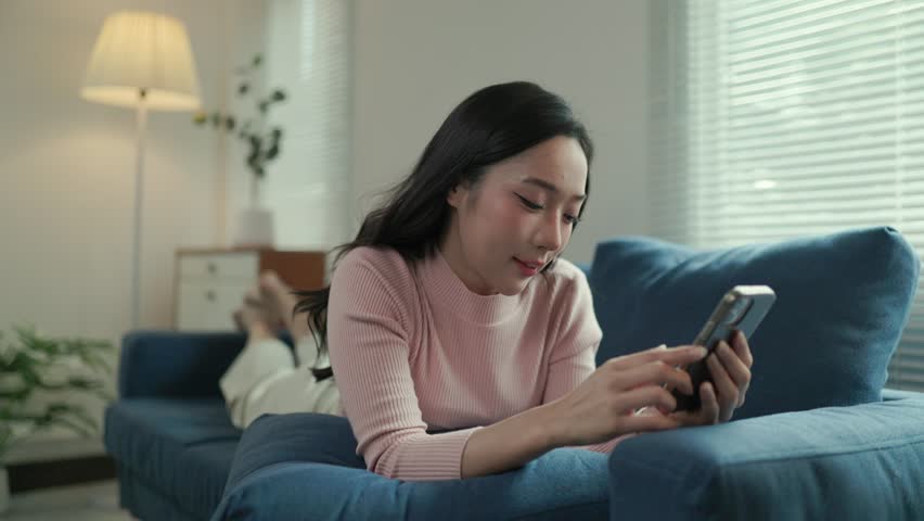 Relaxed young woman lying on cozy blue sofa, navigating social media on smartphone while enjoying peaceful domestic moment during leisure time at home