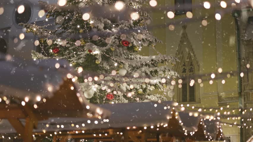 Snowfall creates magical ambiance at holiday market with festive lights and decorated Christmas tree in charming town square. First snow in city for new year holidays