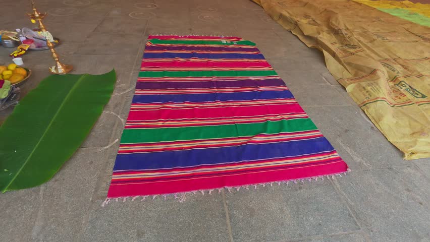 A vibrant handloom "Jamakkalam" carpet with red, green, blue, yellow, and orange stripes and fringed ends lies on a stone floor, reflecting traditional South Indian craftsmanship, used for functions. 