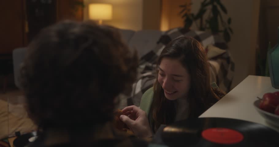 Over the shoulder guy with curly hair a feeds his girlfriend small pieces of pastry during cozy romantic evening with vinyl record in apartment at home