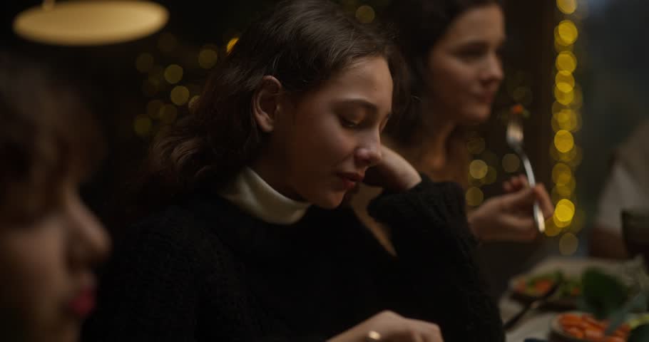 A brunette girl in a black sweater is bored while sitting at the table and having dinner with her family on Christmas evening
