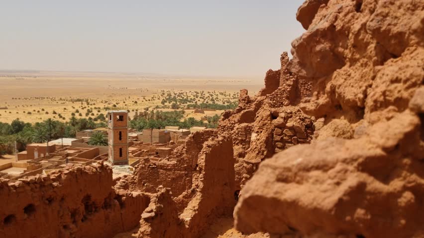 Shot over ancient castle ruins (Ksar) and a nearby traditional village near Timimoun, Algeria, under the blazing sun of a scorching Saharan desert day.