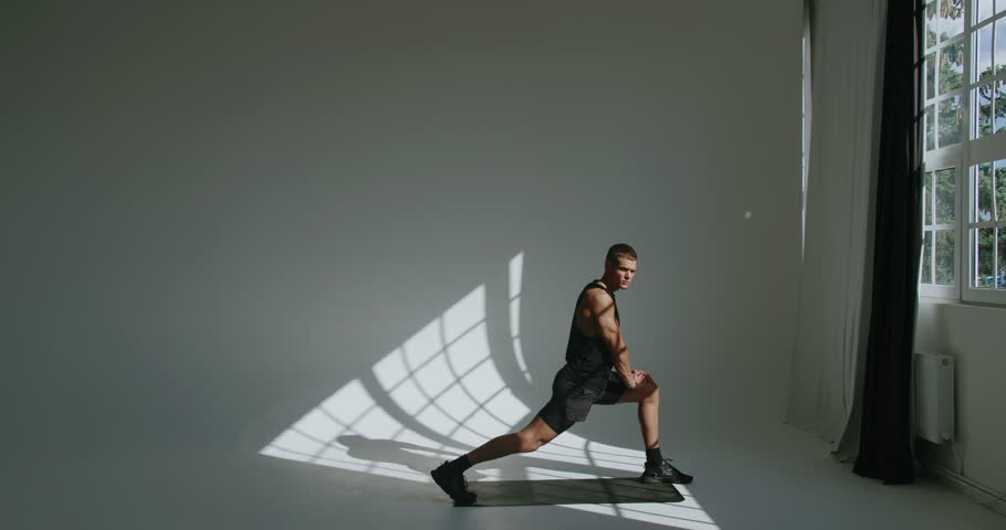 A 23-year-old athletic male stretching in a studio, his figure cast in dramatic light and shadows next to a large window. Young Athletic Man Training in a Bright Studio with Dramatic Shadows