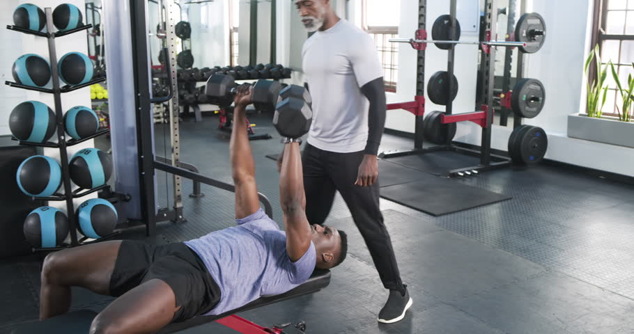 On trainer cue African American man pushing dumbbells on bench while trainer spotting for strength. Strength, fitness, training, gym, workout, motivation, exercise