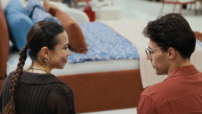 Tilt down of happy young man and woman looking at one another while sitting on sofa in furniture room and discussing collection of textile samples of various colors