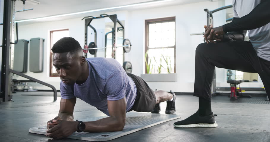 Trainer crouching placing hand on African American male holding plank on mat adjusting posture. Fitness, strength, training, wellness, motivation, discipline, health
