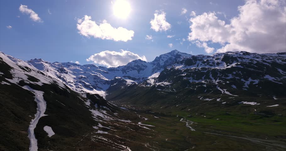 Breathtaking aerial drone footage of a sunlit alpine valley in Montespluga. Shows majestic snow-capped mountain peaks, green slopes, and a dramatic sky. Italian Alps, Lombardy, Italy
