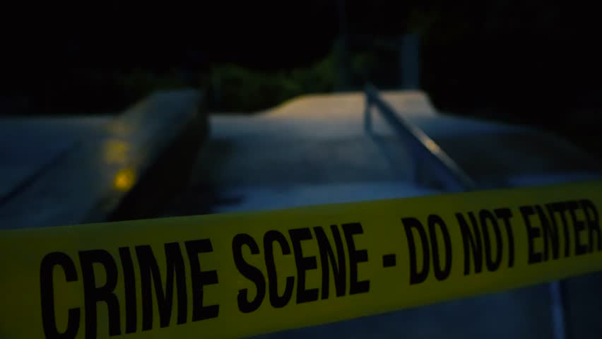 Close up footage glides slowly along fluttering yellow crime scene tape pulled across the skatepark ramps on a moody night. Somber park concrete surface and bushes behind are dimly lit by streetlight