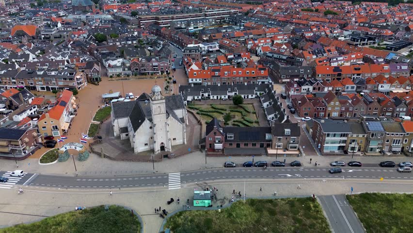 Aerial view of a coastal European town with red-roofed houses, a central church, and a geometric garden. A wide road with cars and a bike lane runs along the foreground near sand dunes.