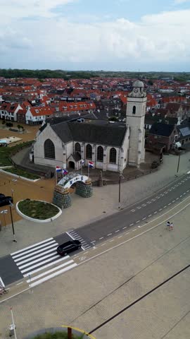 Aerial view of a coastal European town with red-roofed houses, a central church, and a geometric garden. A wide road with cars and a bike lane runs along the foreground near sand dunes.
