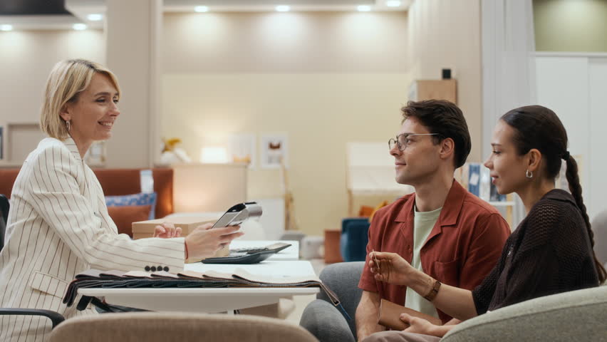 Side view of young couple sitting in front of blond mature female shop assistant with payment terminal while brunette woman paying for purchase by credit card