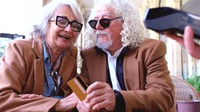 Smiling senior couple makes a credit card payment at an outdoor cafe. They wear stylish coats and sunglasses, enjoying a relaxed moment together in a sunny, casual and urban environment.  - Powered by Shutterstock - Get 15% off with code: PIKWIZARD15