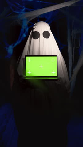 Funny halloween ghost with mockup green screen for advertising or marketing showing computer display, chromakey for advertisement in dark corridor with cobwebs. Happy Halloween