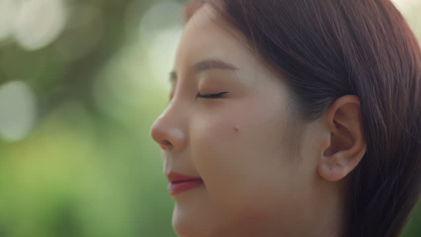 Close-up of peaceful asian woman looking up calmly while enjoying gentle breeze in city park – wellness lifestyle, good health and positive mental wellbeing in relaxing urban nature