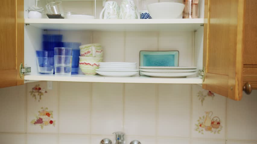 An elderly woman carefully arranges plates and glasses in kitchen cabinets. The warm atmosphere showcases a well-loved home filled with character and personal touches.