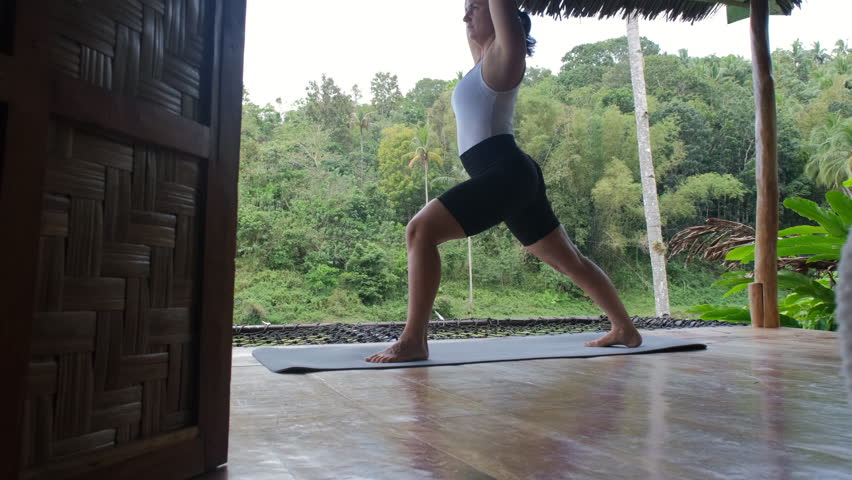 On a terrace of a bamboo hut, a young woman immerses herself in yoga, connecting with nature as she enjoys breathtaking views of rice plantations and vibrant tropical greenery, slow motion