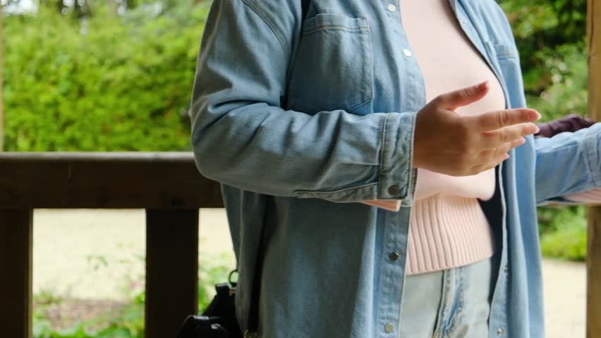 A woman in a blue shirt is standing outside with her hands on her hips. She is holding a camera and she is in a contemplative mood