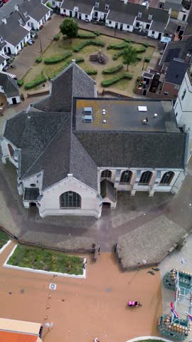 Aerial view of a coastal European town with red-roofed houses, a central church, and a geometric garden. A wide road with cars and a bike lane runs along the foreground near sand dunes.