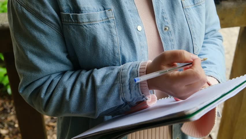 A person is holding a pen and writing in a notebook. The person is wearing a blue jacket and a pink shirt. The notebook is open to a page with writing on it. Scene is focused and determined