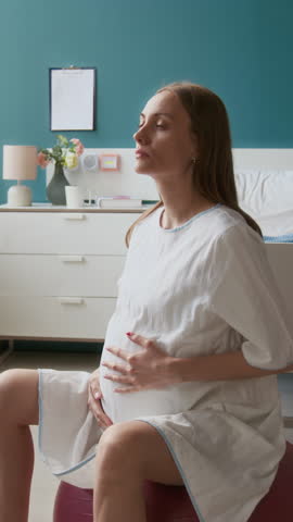 Medium full vertical shot of pregnant young Caucasian woman bouncing on birthing ball in maternity ward and breathing rhythmically to ease intense pain during labor contractions