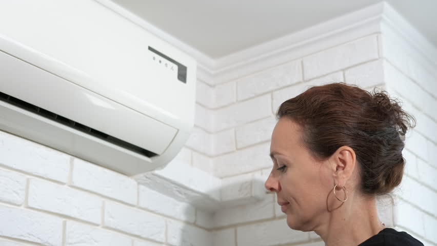 Lady by air conditioner enjoys the cool air. A happy female hold the remote controller and enjoy the cool air from conditioner indoor