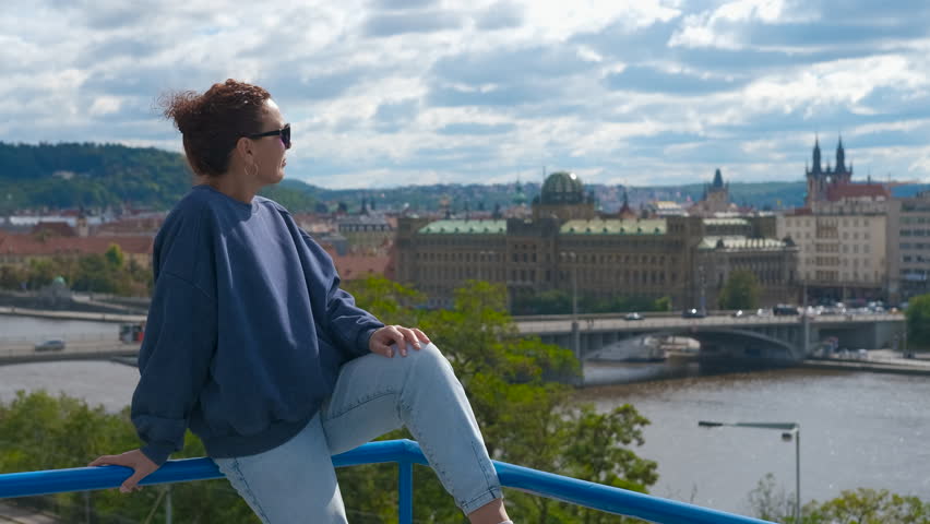 A tourist enjoys the panorama of Prague. Young woman sitting on blue railing, overlooking vltava river and historical prague cityscape, wearing sunglasses and casual attire, enjoying urban panorama