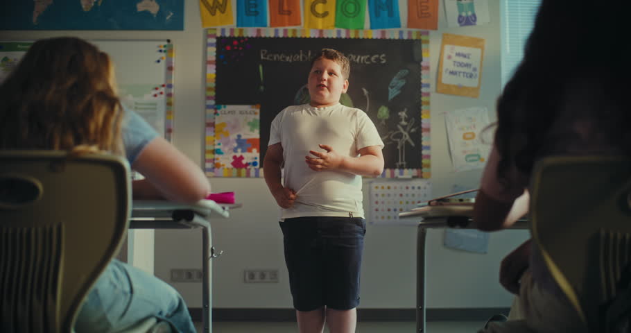 Shy Primary School Boy Showcasing Knowledge of Ecology in Front of Class Full of Kids in Modern Classroom. Female Teacher Supporting Young Student During Presentation. STEM Education. Slow Motion.