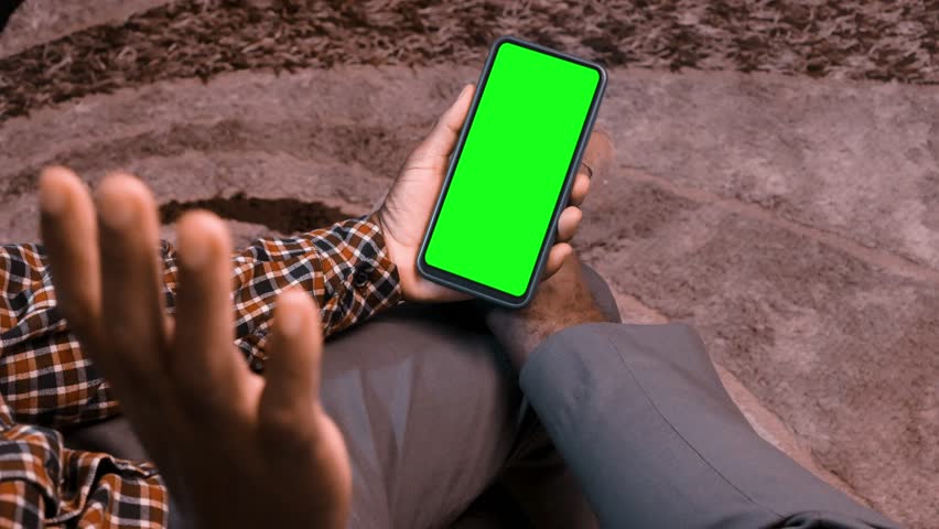 Black African male man sitting in swivel chair watching phone with green screen. Person  waving hands while making video call in room.