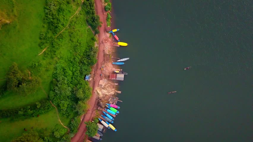 Slow descending aerial view shows colorful wooden boats docked along a rural shoreline of Lake Bunyonyi, Uganda, bordered by green slopes, small structures, and scattered dugout canoes offshore.