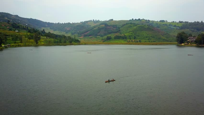 Drone orbiting a traditional dugout canoe gliding over Lake Bunyonyi, Uganda, with lush terraced hills and scattered trees forming a peaceful natural and cultural backdrop.