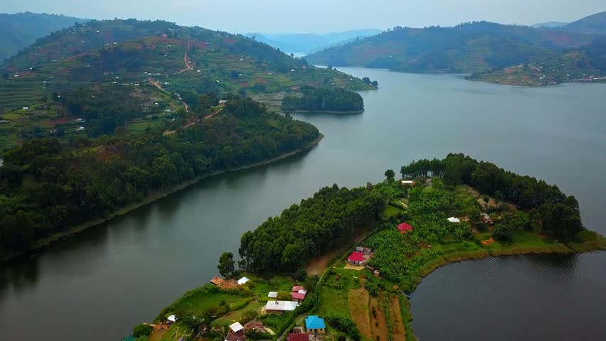 Drone view over Lake Bunyonyi in southwestern Uganda showing green peninsula with cultivated plots and scattered houses in the foreground, and forested terraced hills surrounding the calm lake waters.
