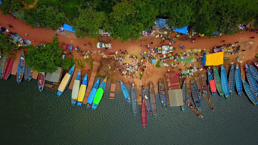 Top-down aerial view of a vibrant weekly market along the red-soil shore of Lake Bunyonyi, Uganda, with docked canoes and colorful tents nestled against dense greenery and forest edge.