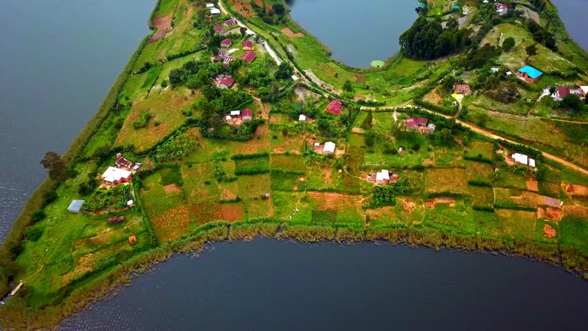 Drone footage shows a curving mainland peninsula on Lake Bunyonyi, Uganda, featuring scattered homes, terraced fields, forested shores, and calm reflective waters between narrow land inlets.