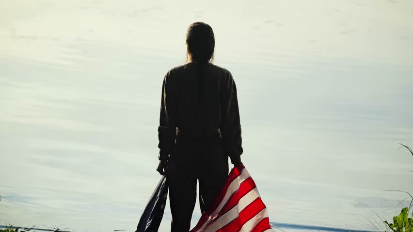 Contemplative woman wrapped in US flag by calm river during soft sunrise light